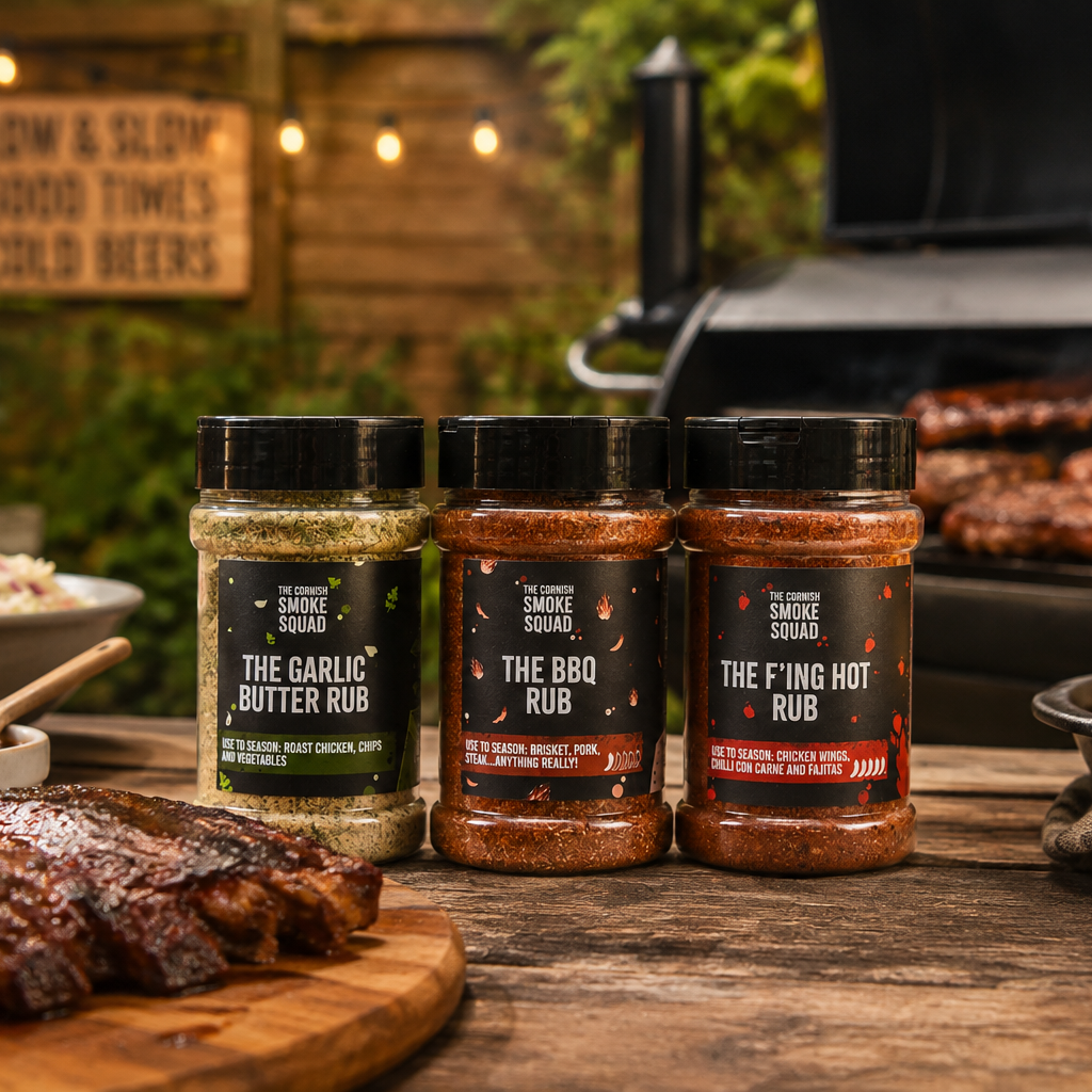 Three rubs from The Cornish Smoke Squad lined up on a wooden table with grilled ribs, corn, and coleslaw in a rustic outdoor backyard setting.