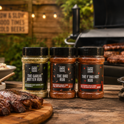 Three rubs from The Cornish Smoke Squad lined up on a wooden table with grilled ribs, corn, and coleslaw in a rustic outdoor backyard setting.